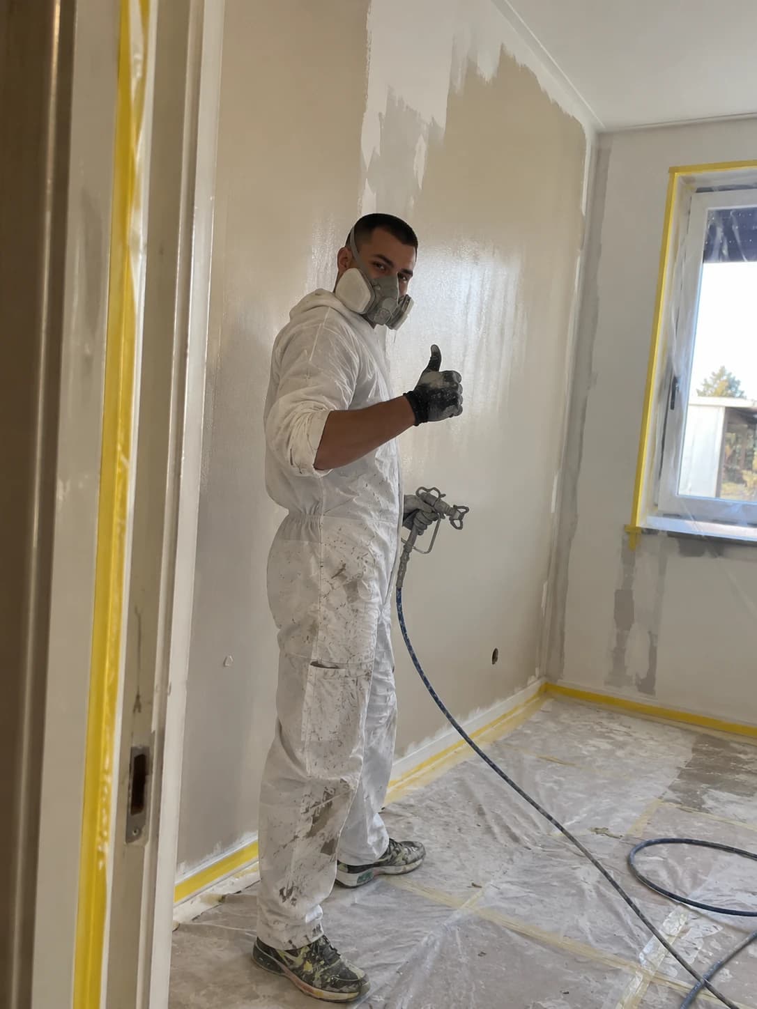 Painter in white overalls and respirator spraying a room wall with latex